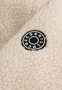 Fleecowa tkanina w kolorze kremowym z teksturowaną powierzchnią. Czarna okrągła metka z białym napisem, oznaczona "OCCODRIL".