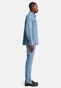 Camicia di denim azzurro chiaro, maniche lunghe con polsini a bottoni, tasca frontale; abbinata a jeans azzurri chiari e mocassini neri.