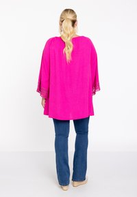 Tunique fuchsia avec des manches texturées, coupe décontractée et bord en dentelle complexe. Associée à un jean bleu évasé et des espadrilles de couleur claire.