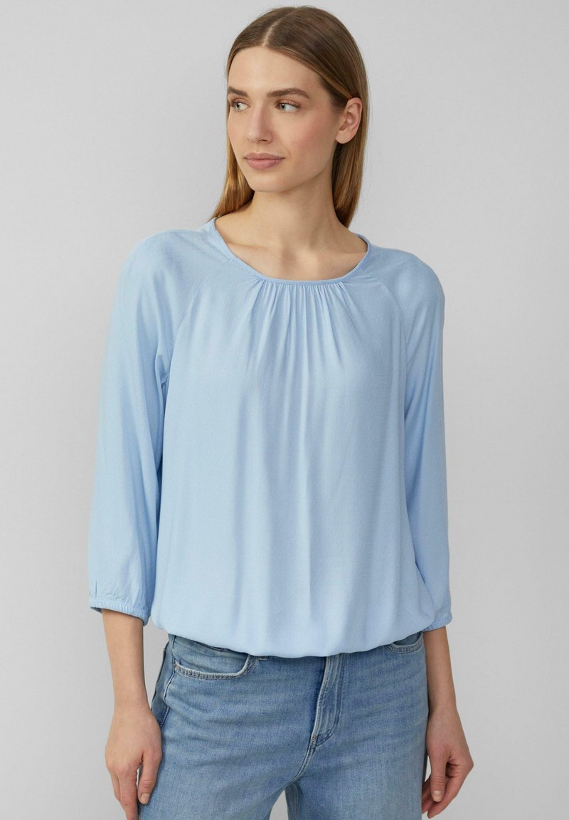 Helles blaues Blusenshirt mit drei Viertel-Raglanärmeln, gerafftem Ausschnitt und lockerer Passform, kombiniert mit Jeans. Weiche, glatte Textur.
