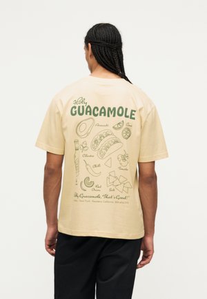 Osoba z zaplecionymi włosami, nosząca beżowy t-shirt z zielonym napisem „Holy Guacamole” oraz ilustracje składników tacos na plecach.