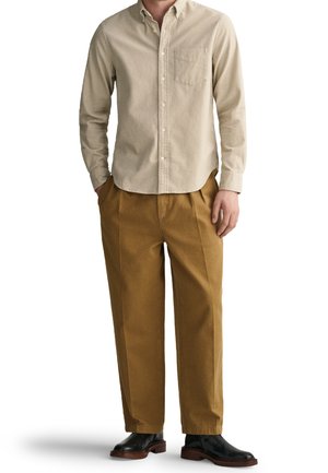 Camisa de botón color beige claro con un bolsillo en el pecho, combinada con pantalones marrones de corte holgado y botas de cuero negras.
