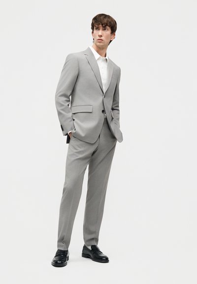 Νεαρός άνδρας που φοράει ανοιχτό γκρι κοστούμι με ταίριαστα παντελόνια, λευκό πουκάμισο και μαύρα δερμάτινα παπούτσια, στέκεται με τα χέρια στις τσέπες.