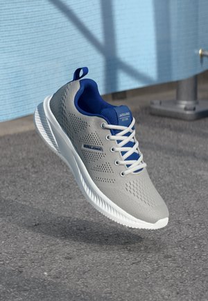 Lys grå og blå Jack & Jones sneakers med hvide snørebånd, svævende over en tekstureret asfaltoverflade i udendørs omgivelser.