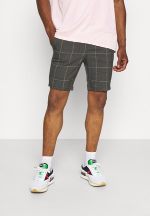 Mann trägt graue karierte Shorts, weiße Socken, weiße Turnschuhe mit schwarzen, grünen und roten Akzenten sowie ein hellrosa Hemd, steht mit entspannten Händen.