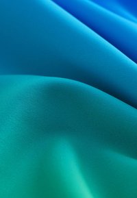 Tessuto strutturato con sfumature degradate di blu e verde teal, che mostra una finitura liscia e morbida con un sottile splendore e onde di varie profondità.
