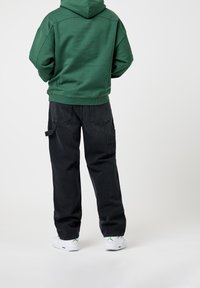 Persona vestida con una sudadera verde, pantalones cargo negros holgados y zapatillas blancas con un logo verde, de pie frente a un fondo blanco.