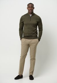 Dunkelgrüner Zip-Halspullover kombiniert mit beigen Stoffhosen und dunkelbraunen Slippern, mit einem taillierten Schnitt und glatter Textur.