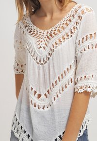 Blouse blanche en tissu lâchement tissé avec des motifs en crochet au niveau du col, des manches et de l'ourlet, portée avec un jean bleu clair.