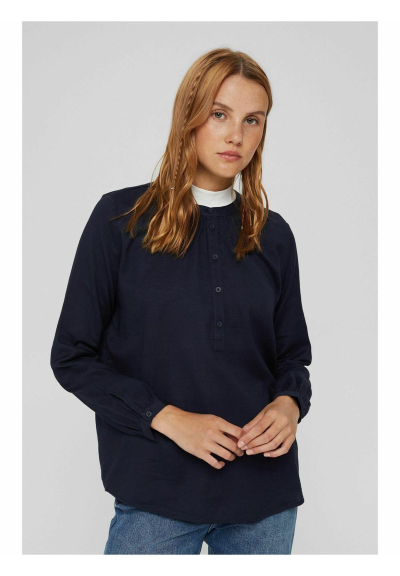 Junge Frau mit hellbraunem Haar in lockeren Zöpfen, die eine navyblaue, knöpfbare Bluse und blue Jeans trägt, steht vor einem einfarbigen Hintergrund.