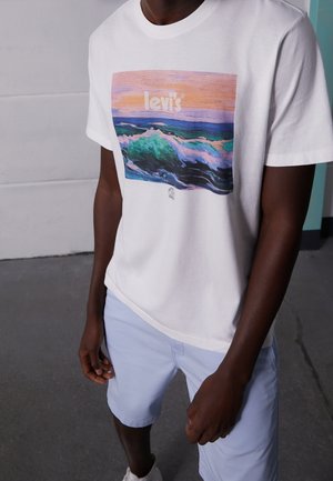 Hombre con camiseta blanca de Levi's con gráfico de ola oceánica y pantalones cortos azul claro, de pie en el interior sobre un suelo gris.