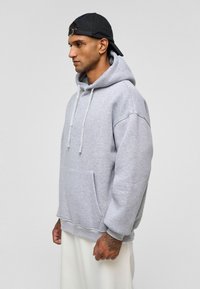 Grijze hoodie met een grote voorzak, trekkoorden en een losse pasvorm. Beschikt over een zachte textuur en geribbelde manchetten. Zwarte pet achterstevoren gedragen.