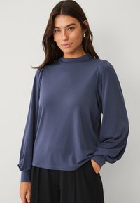 Marineblauwe top met lange mouwen in een gladde stof, met een hoge halslijn, een relaxte pasvorm en gerimpelde mouwdetails. Gecombineerd met zwarte onderkleding.