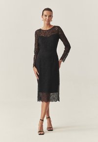Rochie neagră din dantelă cu mâneci lungi, cu modele florale complicate, corset strâns și tiv până la genunchi. Purta cu sandale negre cu toc.