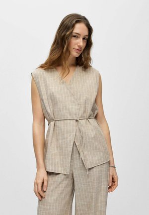 Femme aux longs cheveux ondulés portant un top beige sans manches à fines rayures, noué avec une ceinture, et un pantalon large assorti, debout devant un fond uni.