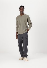 Langärmliger, hellgrüner, strukturierten Pullover kombiniert mit dunkelgrauen Cargo-Hosen mit Taschen. Die Schuhe sind beige mit einer robusten Sohle.