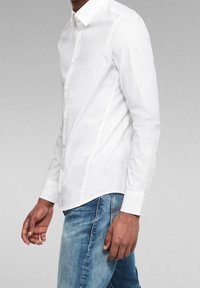 Valkoinen pitkähihaisen napillinen paita, jossa on tavallinen kaulus, valmistettu sileästä kankaasta, yhdistettynä sinisiin farkkuihin, joissa on suora istuvuus.