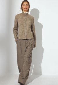 Cardigan en tricot beige avec un col haut et une fermeture à boutons, associé à un pantalon ample couleur brun clair. Design simple et texturé.