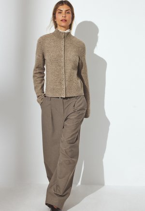 Beige knitted cardigan with a high collar and button closure, paired with loose, light brown trousers. Simple, textured design.