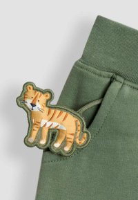 Olivenfarvet stof med et broderet, tegneserieagtigt tigerpatch i gul og orange, med detaljer som striber og blå øjne.