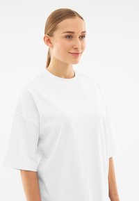 Bílé oversized tričko s krátkým rukávem vyrobené z bavlny, s kulatým výstřihem a volným střihem, předvedeno na modelce s rovnými vlasy.