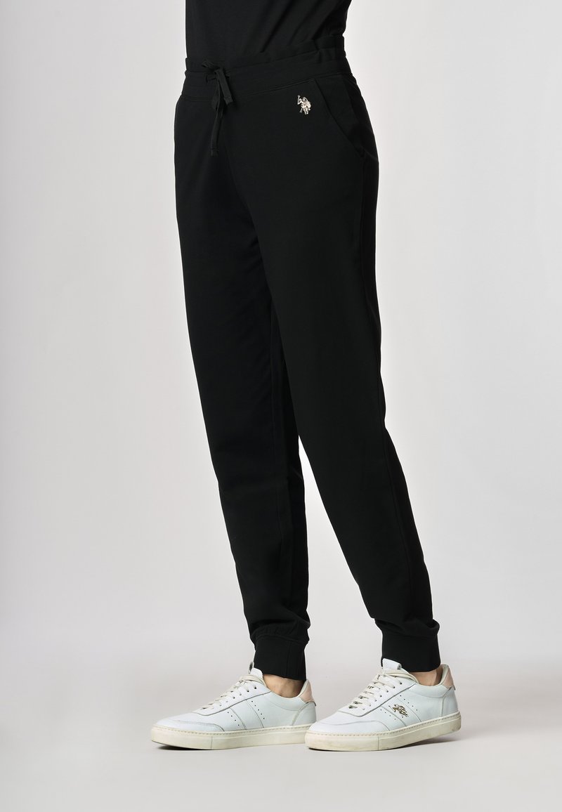 Persona che indossa pantaloni jogging neri con polsini elastici e sneaker bianche, in piedi davanti a uno sfondo chiaro e semplice.