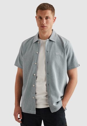 Jonge man draagt een lichtgrijs, kort mouwen, knoop overhemd over een wit T-shirt en zwarte broek, staat met één hand in zijn zak.