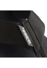 Fekete papucs texturált talppal és szőtt szövetpánttal. Tartalmaz egy apró szürke logótagot, amelyen a "havaianas" felirat szerepel a pánton.
