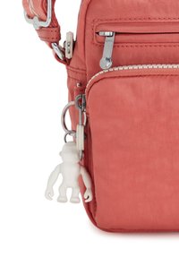 Korallfärgad tygväska med flera zippered fack, silverfärgad hårdvara och en vit apa charm fäst vid en dragkedja. Strukturerad yta.