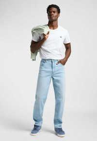 Vaqueros de denim azul claro de corte recto, combinados con una camiseta blanca de cuello redondo. Un suéter verde menta colgado sobre un hombro.