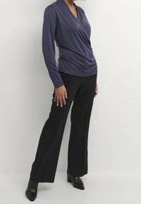 Kvinna klädd i en mörkblå långärmad inlindad topp, svarta vida byxor och svarta klackiga ankelstövlar stående mot en enfärgad bakgrund.