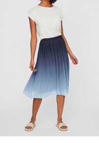 Vrouw in een witte korte mouw top en een geplisseerde midi-jurk met een blauwe ombré-gradient, gecombineerd met beige instap-sandalen.