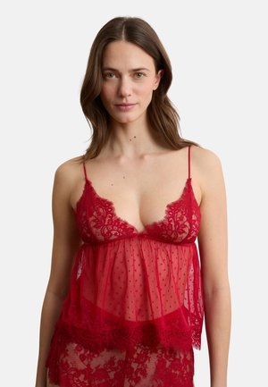 Donna che indossa lingerie in pizzo trasparente rosso con motivi floreali e sottili spalline, rivolta verso l'obiettivo su uno sfondo chiaro uniforme.