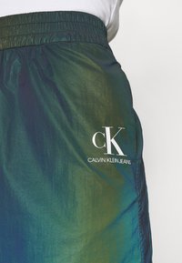 Nahaufnahme von dunkelgrünen Calvin Klein Jeans mit elastischem Bund und weißem "cK Calvin Klein Jeans"-Logo auf dem Oberschenkel.