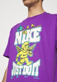 Lila bomullströja med en grafik av en gul cherub och en ros, med texten "NIKE JUST DO IT" i fet vit och blå konturer.