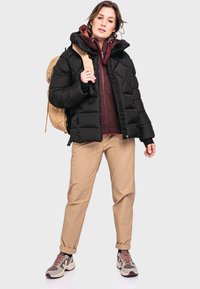 Schwarze Pufferjacke mit Kapuze, bordeauxrotes, mehrlagiges Fleece, beige Hose und graue Sneakers mit rosa Schnürsenkeln, sowie einen beigen Rucksack.