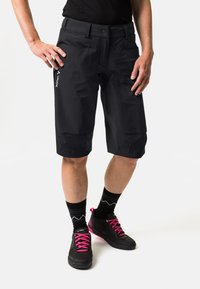 Schwarze Shorts aus leichtem Material mit Knopfverschluss, zwei Fronttaschen und einem dezenten Logo. Kombiniert mit schwarzen Schuhen und pinken Schnürsenkeln.