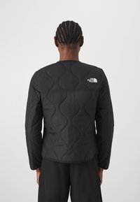 Černá prošívaná bunda s dlouhými rukávy, s kulatým lemem a bílým logem na horní části zad. Materiál se zdá být hladký a lehký.