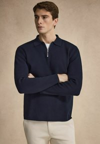 Mörkblå pullover med halvdragkedja vid kragen, ribbad struktur, långa ärmar och rak nederkant, kombinerad med off-white byxor.