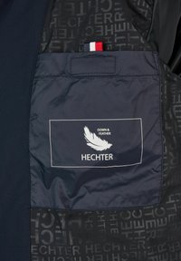 Marineblå interiør i en jakke med en lomme, der har en hvid etiket, prægede fjerlogo og "HECHTER"-tekst i et mørkt gentaget mønster.
