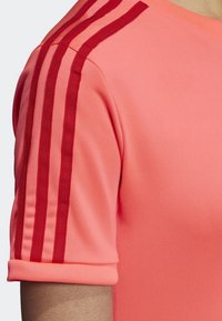 Krátký rukáv atletického trička v jasně korálové barvě, s třemi vertikálními červenými pruhy na každém rameni a žebrovaným manžetovým lemem. Hladká, pružná látka.