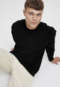Bluză neagră cu mâneci lungi dintr-o textură moale, cu decolteu rotund, asortată cu pantaloni din corduroy crem, așezată pe un fundal neutru.