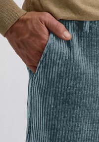 Teal corduroy broek met verticale ribbels, voorzien van een zijzak. Een hand is in de zak geplaatst, waardoor de textuur en kleur worden benadrukt.