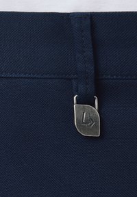 Granatowy materiał w pasie spodni z srebrnym metalowym zapięciem z logo. Tekstura jest gładka i mocno tkana.