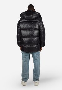 Muž v černé péřové bundě s kapucí, světlých modrých cargo džínách a bílých teniskách, stojící zády k bílé pozadí.