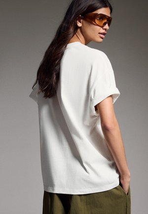 Ženska z dolgimi temnimi lasmi nosi velike sončna očala z rjavim odtenkom, ohlapno belo majico s povihanimi rokavi in olivno zelene hlače ter gleda v stran.