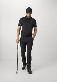 Μαύρο polo γκολφ με ριγέ λεπτομέρειες, σε συνδυασμό με μαύρους στενούς παντελόνες και μαύρα παπούτσια. Η φιγούρα κρατάει μια ράβδο γκολφ, ολοκληρώνοντας την αθλητική εμφάνιση.