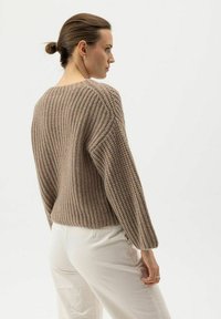 Frau mit einem Dutt, die einen strukturierten braunen Strickpullover und helle Hosen trägt, steht im Profil vor einem schlichten Hintergrund.