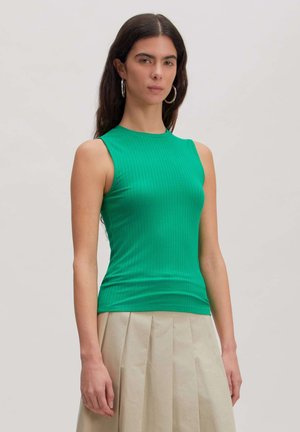 Frau mit langen dunklen Haaren, die silberne Creolen-Ohrringe, ein ärmelloses grünes geripptes Top und einen beigen plissierten Rock vor einem schlichten Hintergrund trägt.