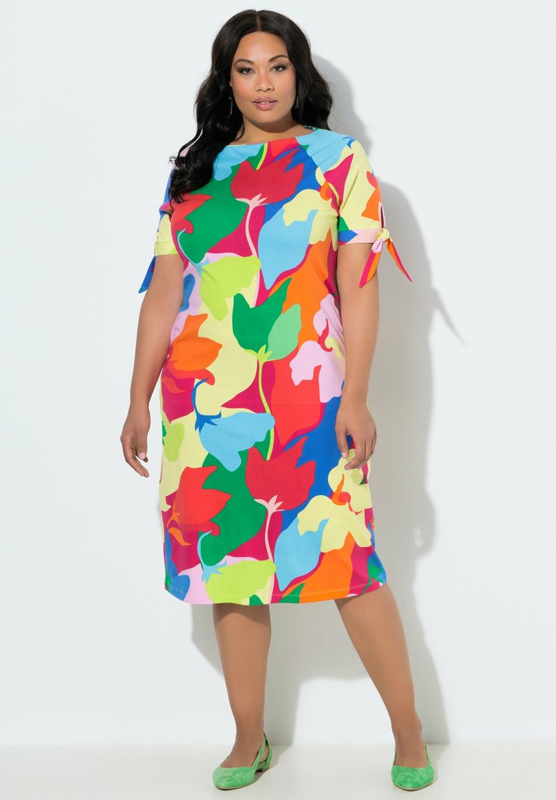 Frau trägt ein knielanges, kurzärmeliges Kleid mit großen, bunten Blattmustern und grüne Schuhe mit niedrigem Absatz, steht vor einer weißen Wand.